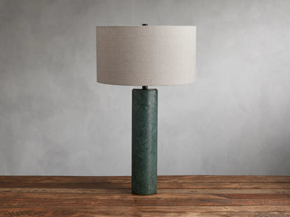 Brixton Table Lamp with a cylindrical green marble base and a large cream fabric lampshade, placed on a wooden surface against a gray wall.