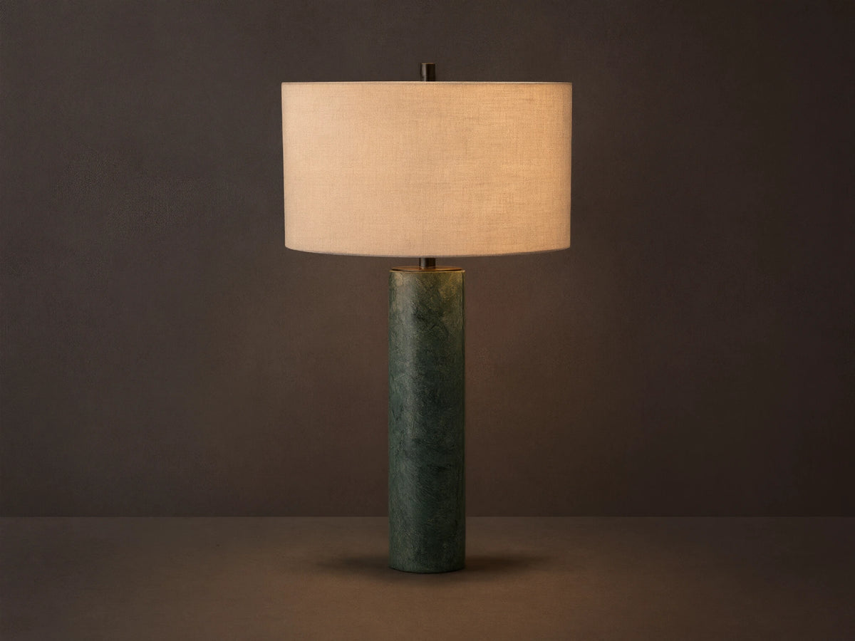 Brixton Table Lamp with a cylindrical green marble base and a large cream fabric lampshade, placed on a wooden surface against a gray wall.