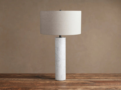 Brixton Table Lamp, a cylindrical white marble base, dark metal neck, and light grey fabric shade on a rustic wood surface.