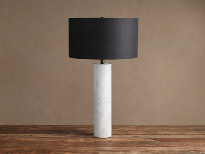 Brixton Table Lamp featuring a tall, cylindrical white marble base with gray veins and a black fabric drum shade, on a wood surface.