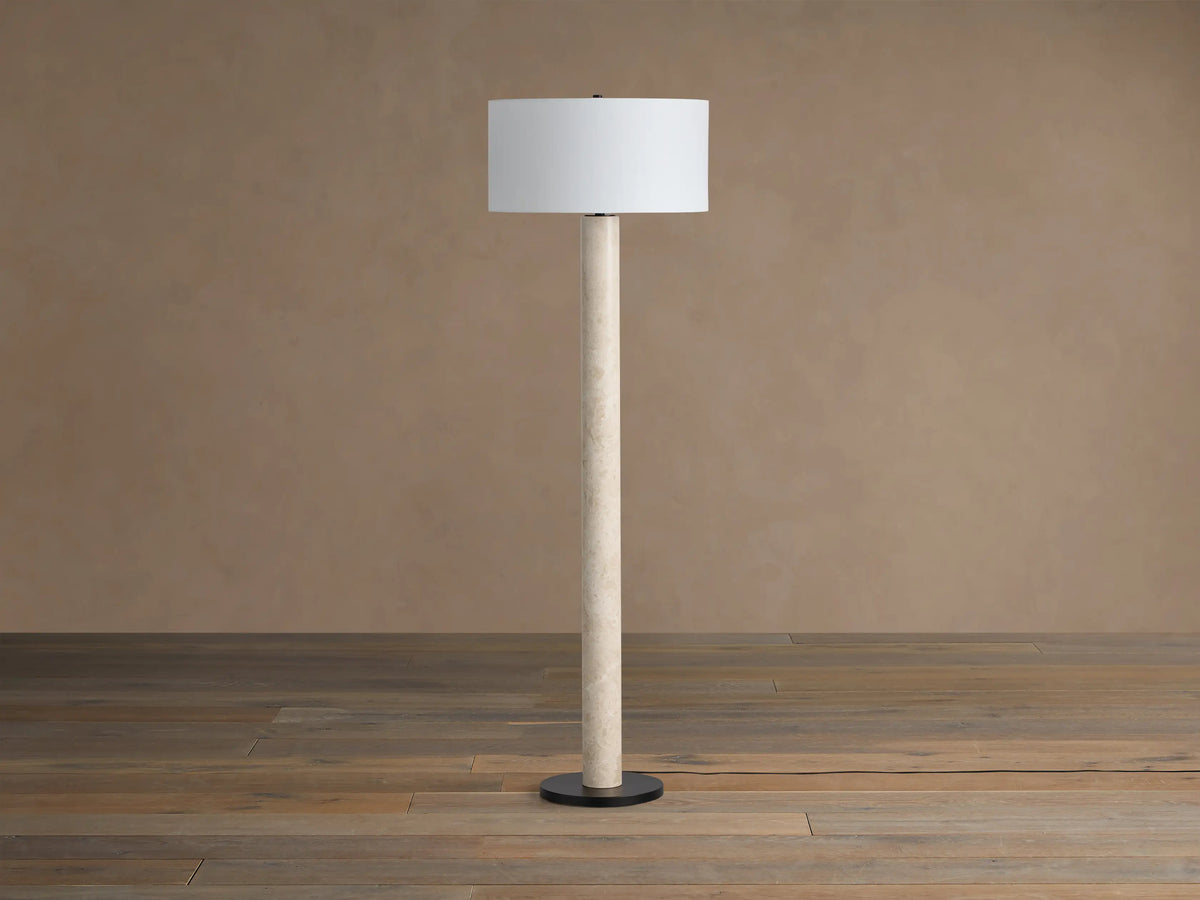 Brixton Floor Lamp with a tall, light beige marble-look stem, white cylindrical shade, and round black base on a wood floor.