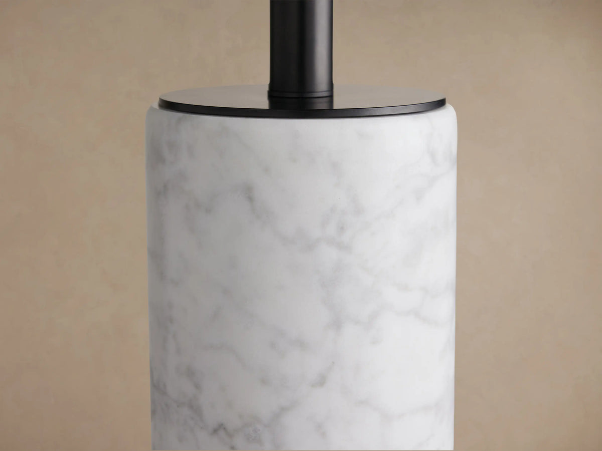 Brixton Floor Lamp with white marble column, light fabric shade, and black base, standing on a wooden floor against a brown wall.