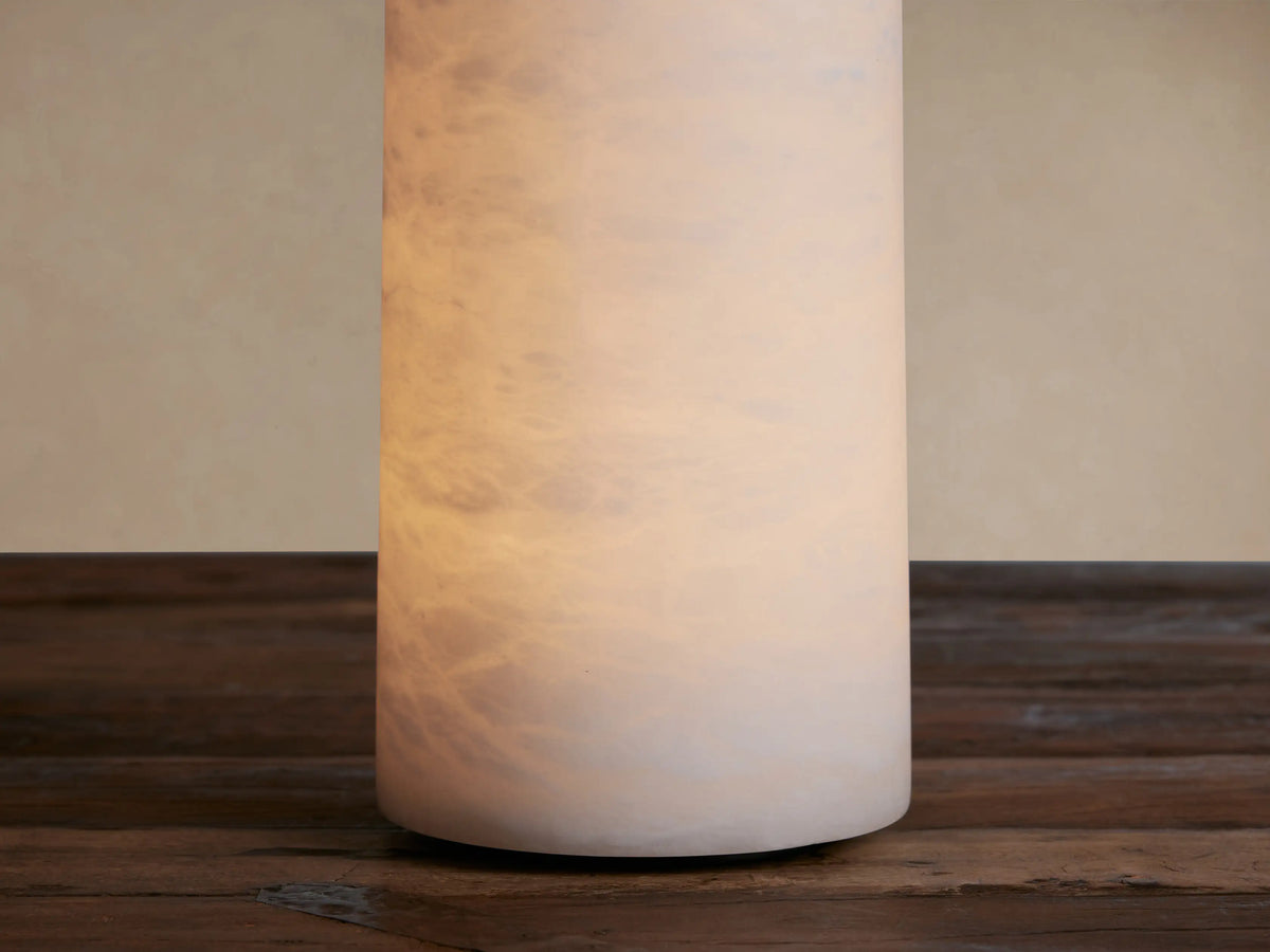 White cylindrical Kelby Outdoor Rechargeable Lamp made of alabaster with natural patterns, standing on a dark wooden surface.