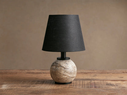 Celia Outdoor Rechargeable Lamp with a spherical cream/grey travertine base, black linen shade, and matte black hardware on a rustic wooden table.