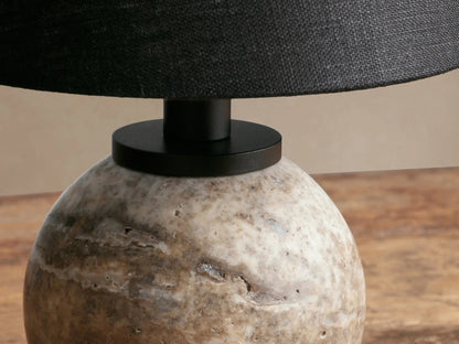 Celia Outdoor Rechargeable Lamp with a rounded, textured beige travertine base and a black conical linen shade on a wood table.