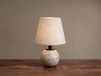 Celia Outdoor Rechargeable Lamp with a round, textured travertine base, matte black hardware, and an illuminated flax linen shade.