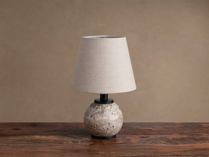 Celia Outdoor Rechargeable Lamp with a round, textured travertine base, matte black hardware, and an illuminated flax linen shade.