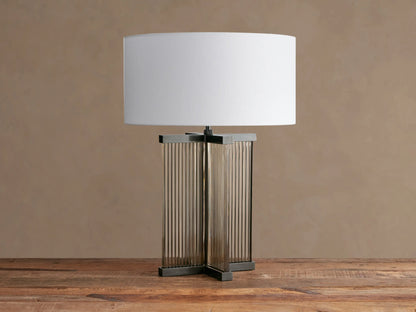 Delsie Table Lamp with an off-white shade, clear fluted glass base with dark bronze framing, on a rustic wood table.