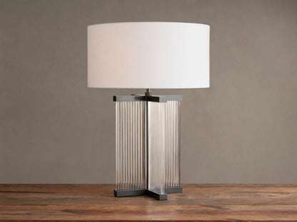 Delsie Table Lamp with a white drum shade over a dark metal base with clear fluted glass panels, on a rustic wooden table.