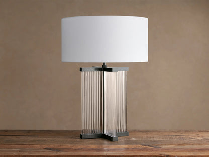 Delsie Table Lamp with a white drum shade over a dark metal base with clear fluted glass panels, on a rustic wooden table.