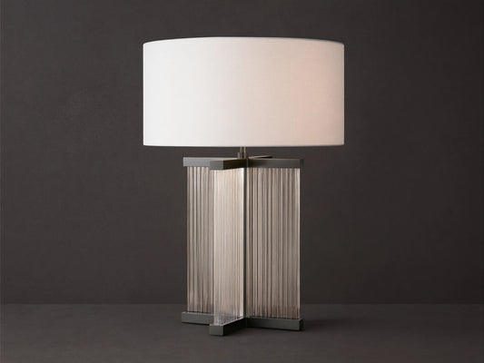 Delsie Table Lamp with a white drum shade over a dark metal base with clear fluted glass panels, on a rustic wooden table.