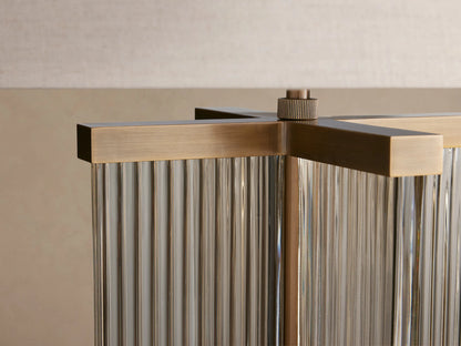 Delsie Table Lamp with a light flax cylindrical shade and a fluted clear glass base with burnished brass accents, on a wooden table.