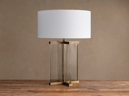 Delsie Table Lamp, white fabric shade atop a fluted glass and burnished brass base, sitting on a rustic wooden surface.