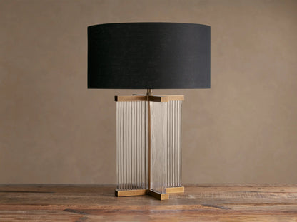 Delsie Table Lamp with a black drum shade, clear fluted glass rectangular base, and burnished brass accents on a wooden table.