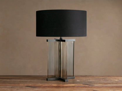 Delsie Table Lamp with a black fabric drum shade, fluted clear glass body, and burnished bronze metal base on a wood table.