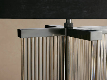 Delsie Table Lamp. Black drum shade atop a fluted glass base with burnished bronze metal, on a rustic wooden table.