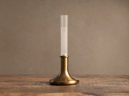 Luella Outdoor Rechargeable Lamp: burnished brass base & clear glass tube with glowing LED, standing on a rustic wood table.