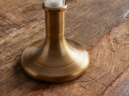 Luella Outdoor Rechargeable Lamp with a burnished brass base & clear glass cylinder on a wooden table, against a brown wall.