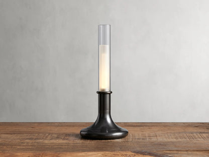 Luella Outdoor Rechargeable Lamp with brushed bronze finish, cylindrical glass top, glowing LED, on wooden table, grey background.