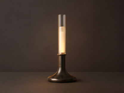 Luella Outdoor Rechargeable Lamp with brushed bronze finish, cylindrical glass top, glowing LED, on wooden table, grey background.