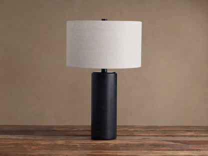 Brixton Table Lamp with a dark, cylindrical marble base & light fabric drum shade on a rustic wooden table.