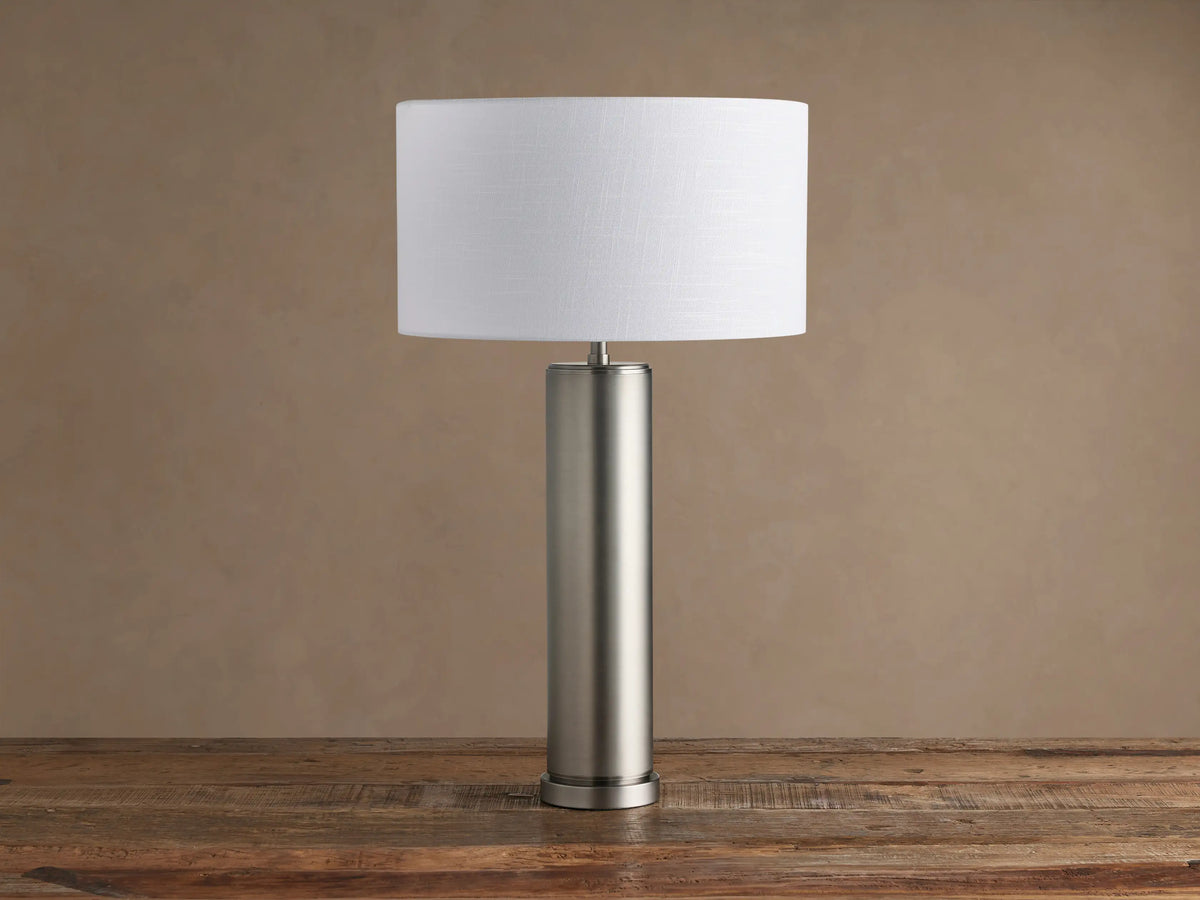 Linden Table Lamp in pewter with cylindrical base, white drum shade, on rustic wooden table against brown wall.