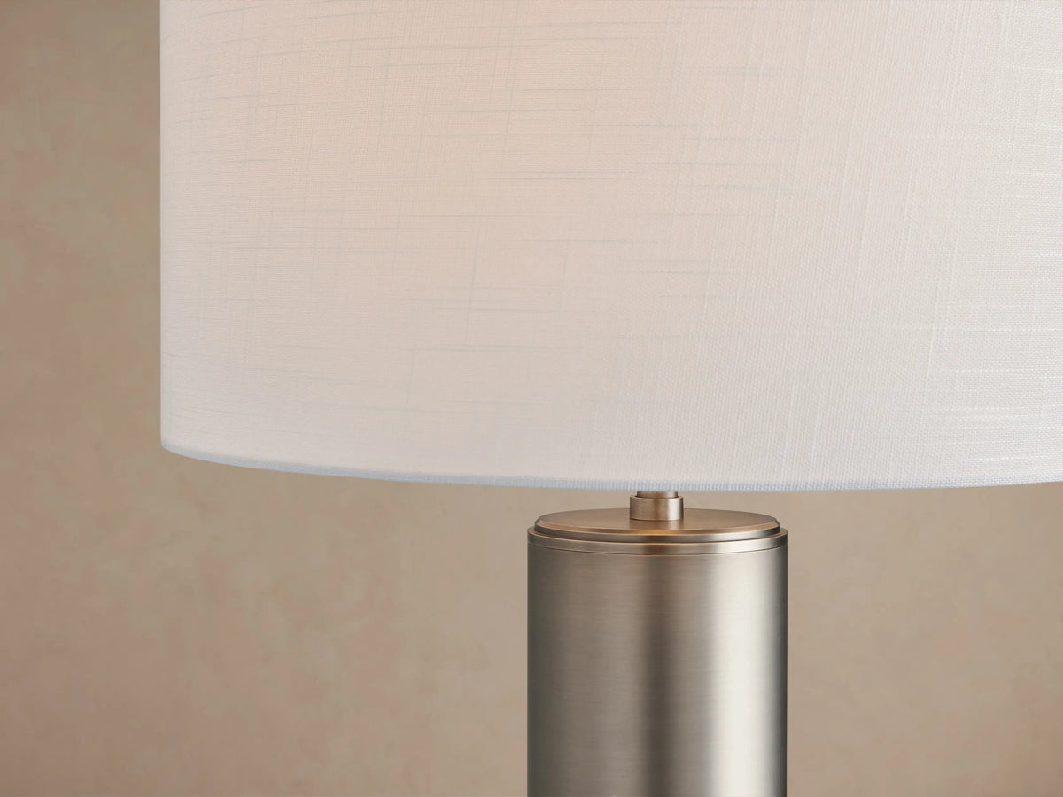 Linden Table Lamp with a cylindrical pewter base, topped by a white linen drum shade, sitting on a rustic wooden table.