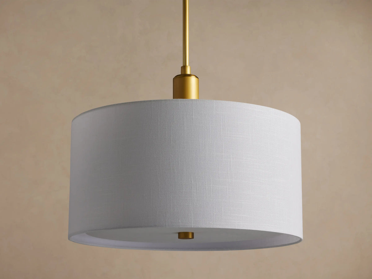 Linden Pendant with a glowing white linen drum shade hanging from a brass rod, seen at a slight angle on a beige background.
