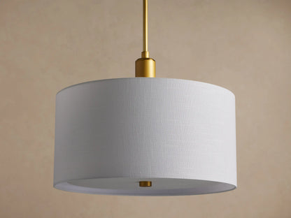 Linden Pendant with a glowing white linen drum shade hanging from a brass rod, seen at a slight angle on a beige background.