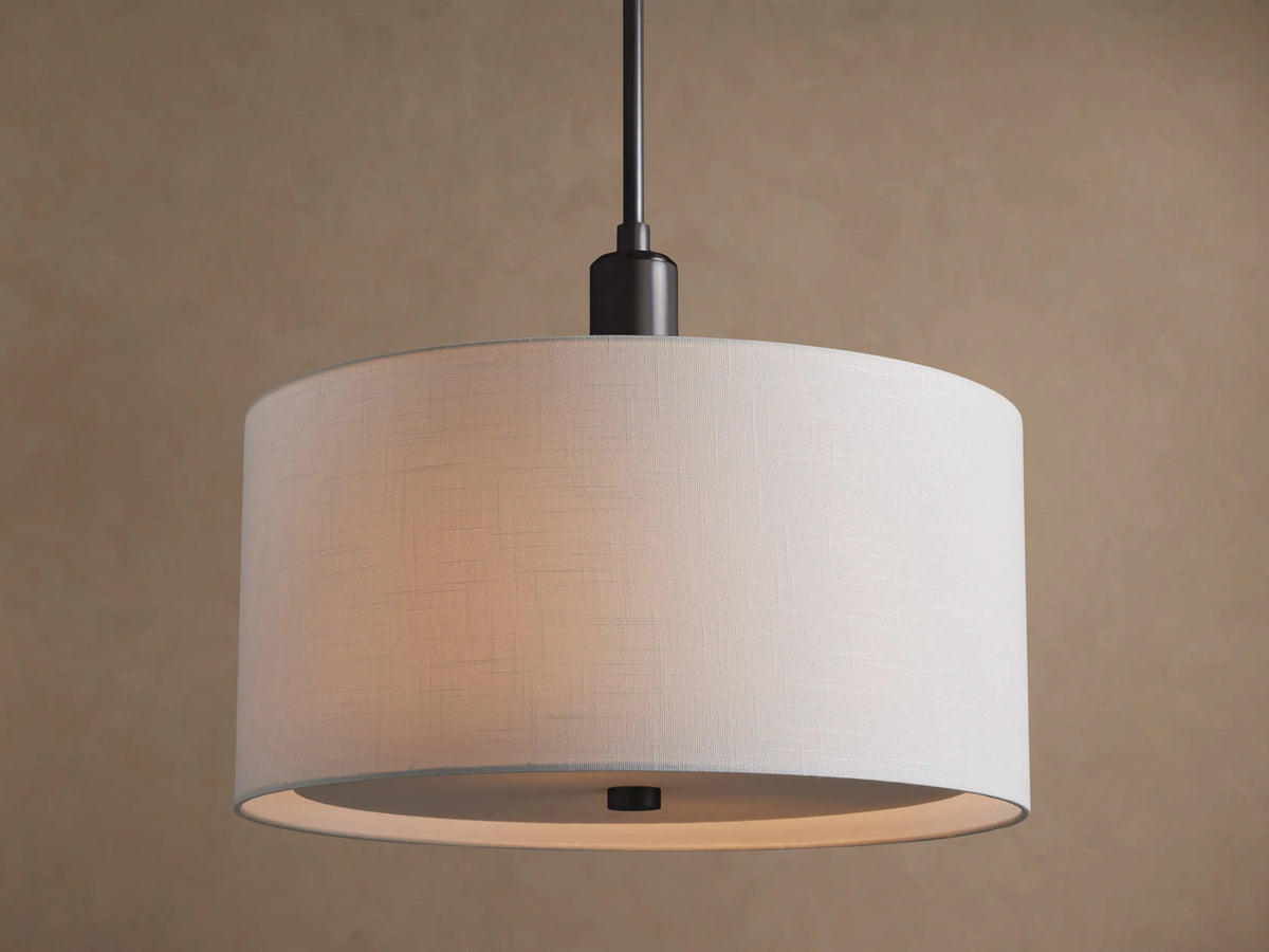 A Linden Pendant hangs, showing its large, round white linen drum shade and dark bronze stem, against a textured brown wall.