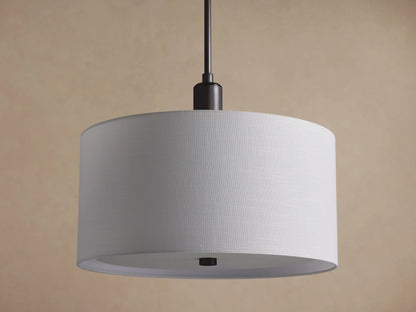 A Linden Pendant hangs, showing its large, round white linen drum shade and dark bronze stem, against a textured brown wall.