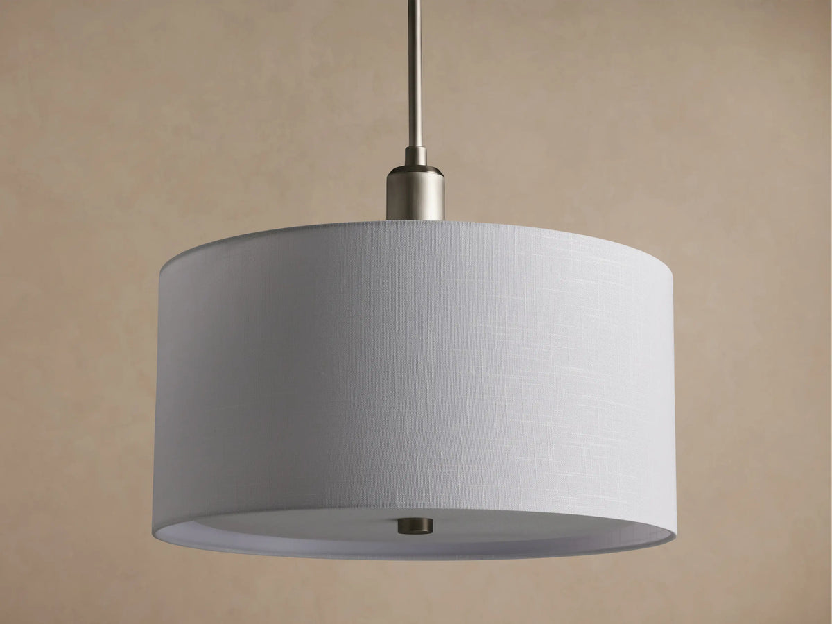 Linden Pendant with cylindrical, off-white linen shade, brushed metallic hardware, glowing softly against a warm wall.