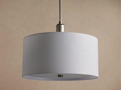 Linden Pendant with cylindrical, off-white linen shade, brushed metallic hardware, glowing softly against a warm wall.