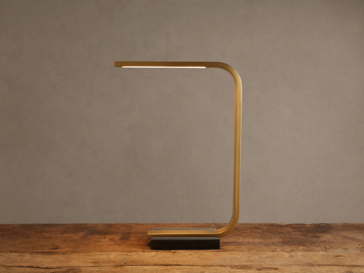 Karson Task Table Lamp: sleek brass, U-shaped lamp with an illuminated LED strip, on a black marble base, resting on a wooden table.