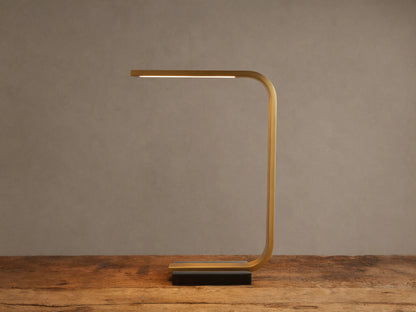 Karson Task Table Lamp: sleek brass, U-shaped lamp with an illuminated LED strip, on a black marble base, resting on a wooden table.