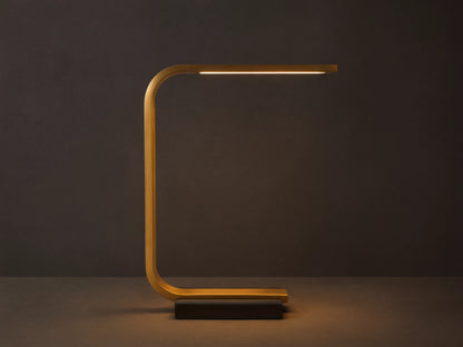 Karson Task Table Lamp: sleek brass, U-shaped lamp with an illuminated LED strip, on a black marble base, resting on a wooden table.