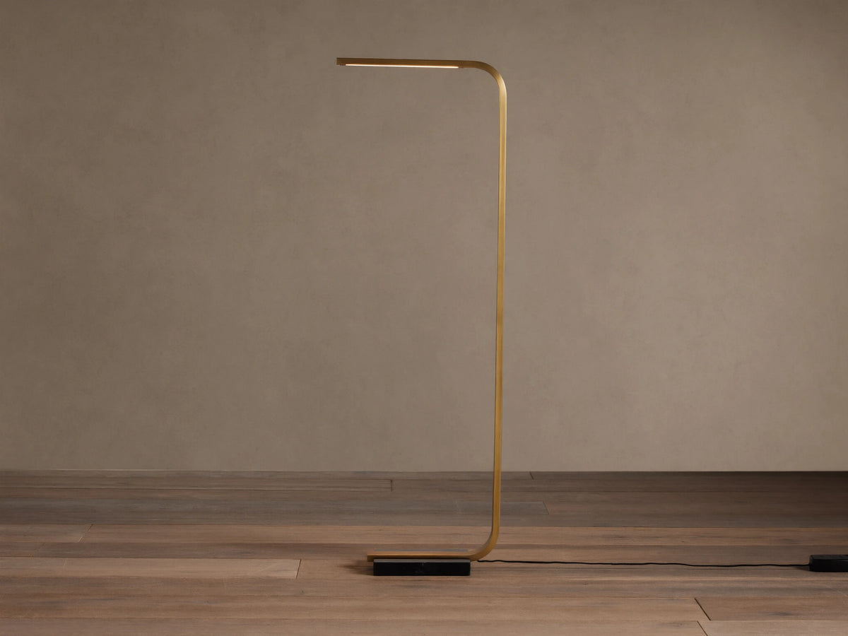 Modern brass Karson Task Floor Lamp with a curved stem, glowing light strip, and black marble base on a wood floor.