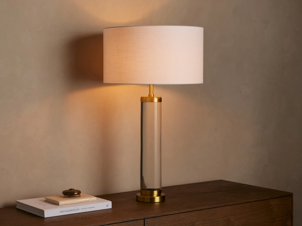 Linden Crystal Table Lamp featuring a clear crystal column, brass accents, and an illuminated white linen shade on a wood table.