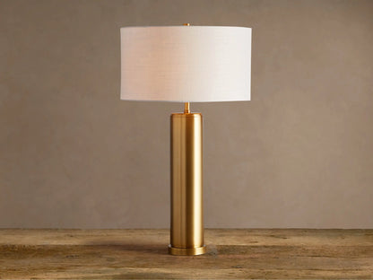 Linden Table Lamp: cylindrical brass-finished iron base, white linen drum shade, on a rustic wood surface.