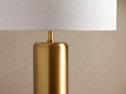 Linden Table Lamp with a polished brass cylindrical base and a white linen drum shade, on a rustic wood table.