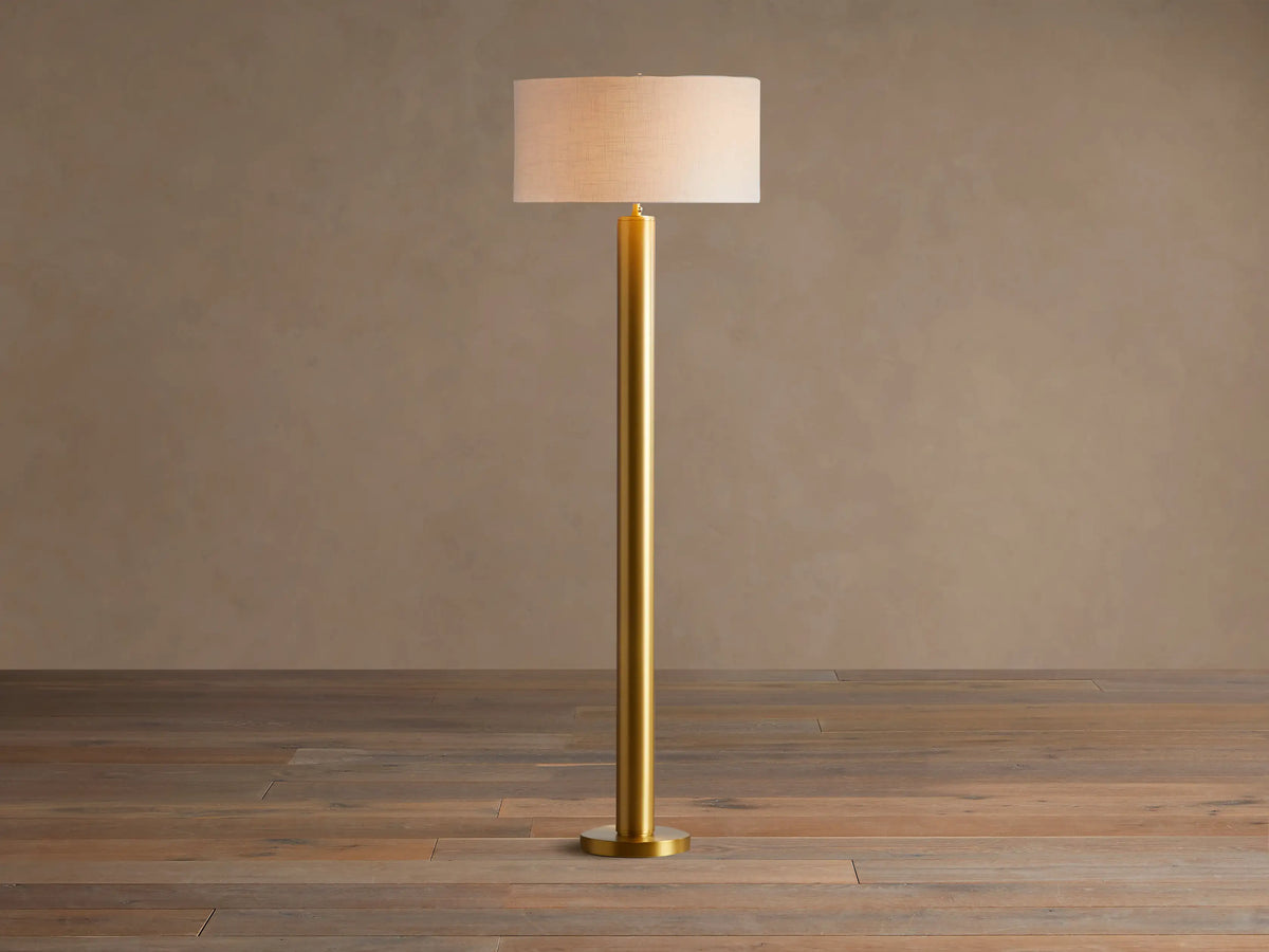 Linden Floor Lamp with a brass-finished pole and round base, topped by a light beige linen drum shade. Standing on a wood floor.
