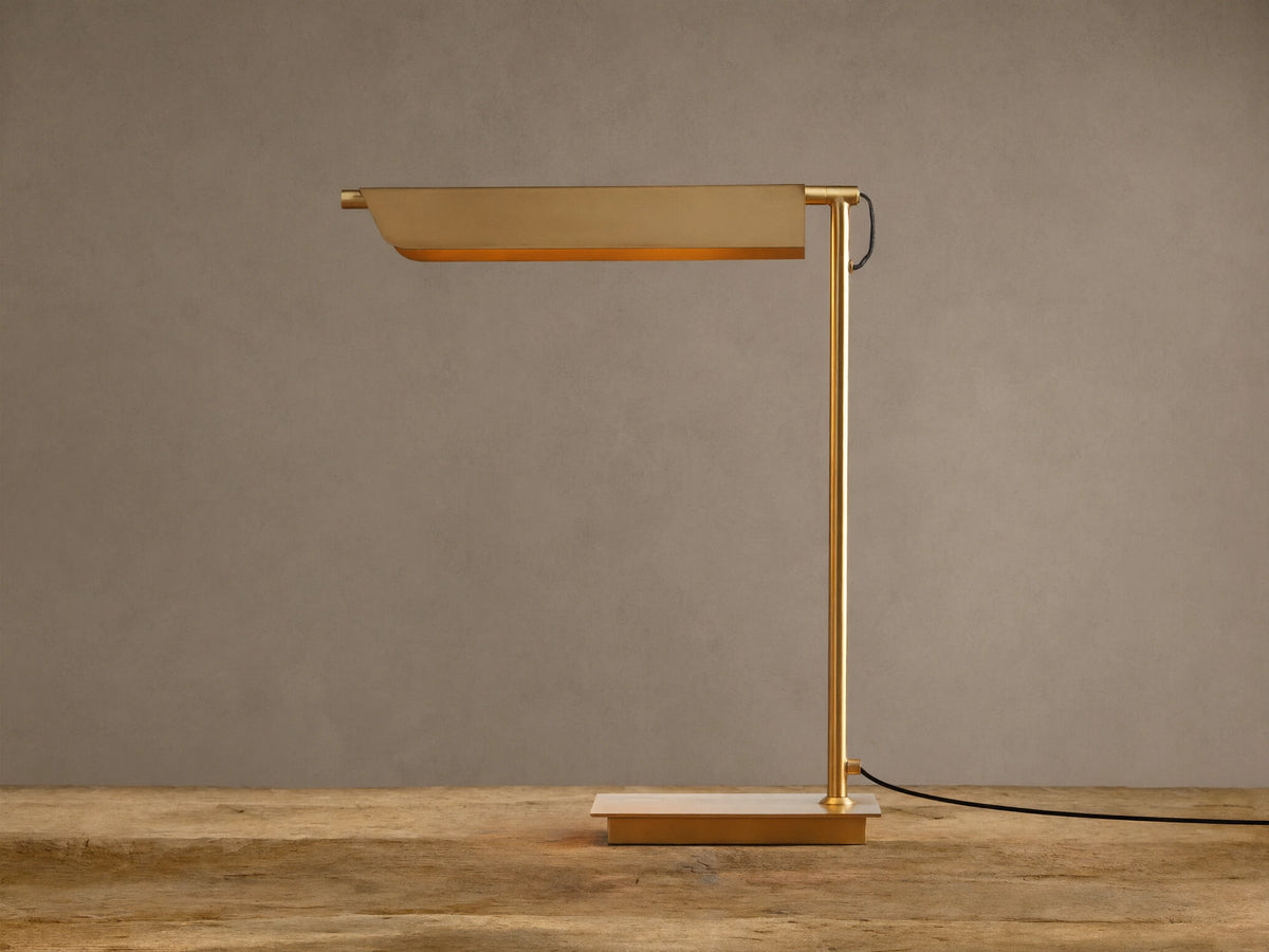 Modern Axle Brass Table Lamp with rectangular shade emitting warm light, slender stand, on rustic wood. Neutral wall background.