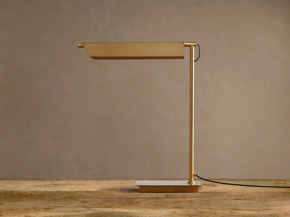 Modern Axle Brass Table Lamp with rectangular shade emitting warm light, slender stand, on rustic wood. Neutral wall background.
