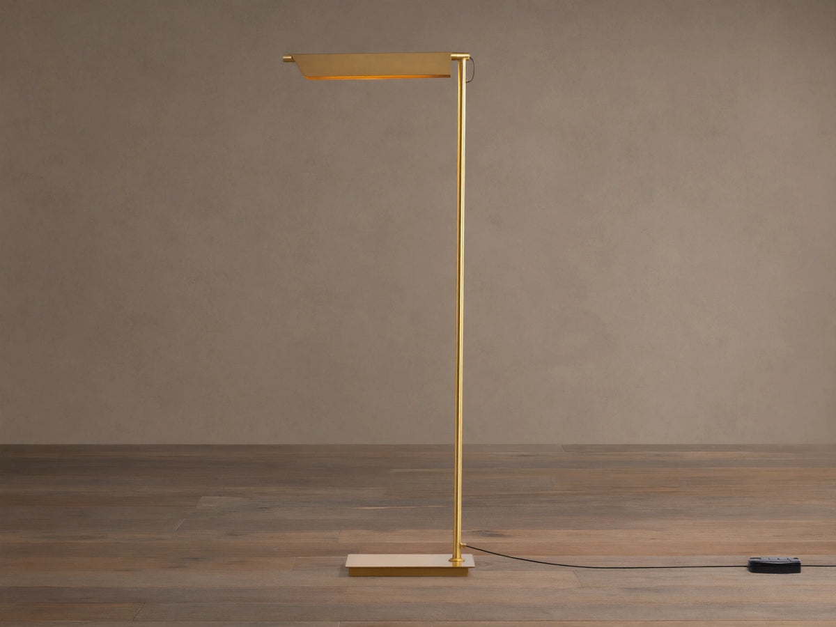 Axle Brass Floor Lamp with a glowing rectangular shade, slim brass pole, square base, and black dimmer on a wooden floor.