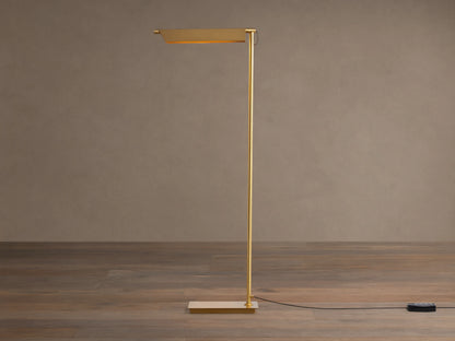 Axle Brass Floor Lamp with a glowing rectangular shade, slim brass pole, square base, and black dimmer on a wooden floor.