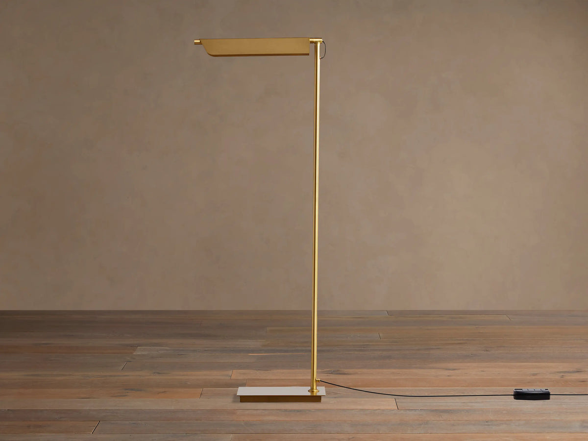 Axle Brass Floor Lamp with a glowing rectangular shade, slim brass pole, square base, and black dimmer on a wooden floor.