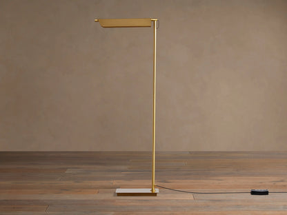 Axle Brass Floor Lamp with a glowing rectangular shade, slim brass pole, square base, and black dimmer on a wooden floor.