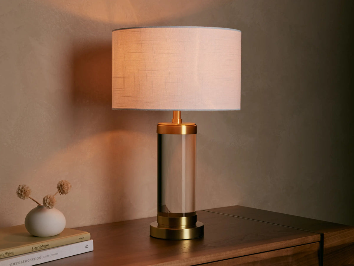 Linden Crystal Table Lamp with clear crystal column, brass accents, and off-white linen shade, glowing on a wooden table.