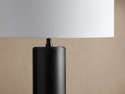 Linden Table Lamp with a dark cylindrical base and a white linen cylindrical shade, sitting on a rustic wooden table.