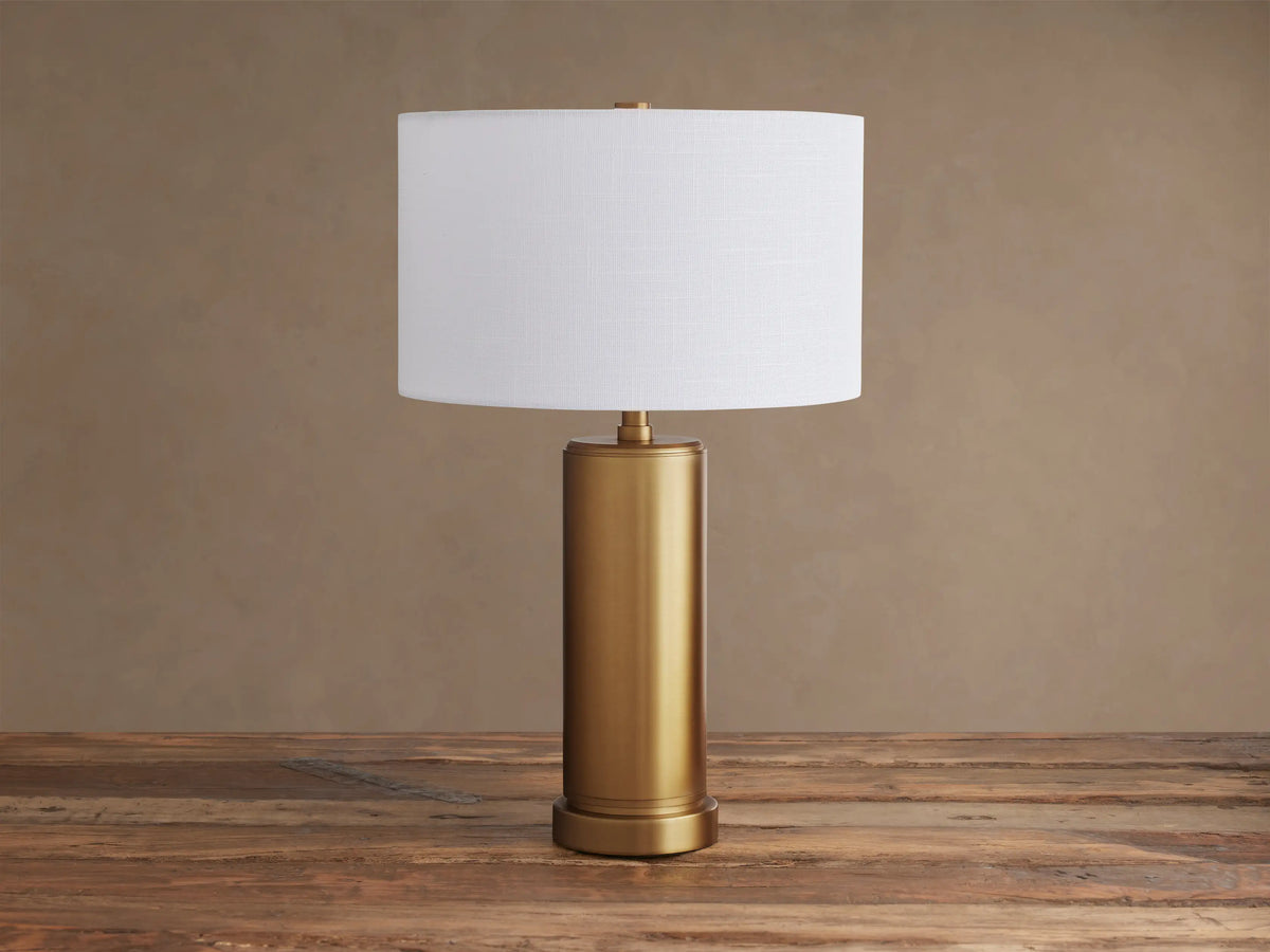 Linden Table Lamp with a brass-finished cylindrical base and a glowing white linen drum shade on a rustic wooden table.